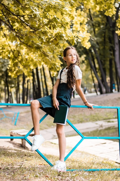 Schoolgirl holding a book and sitting on a fence in a park on a warm day Vertical view