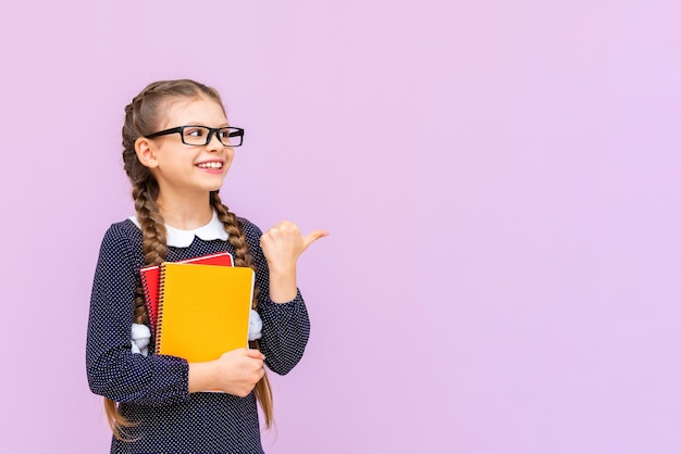 A schoolgirl holds notebooks and points to your advertisement on a pink isolated background Educational courses for secondary school children Copy space