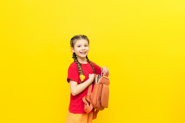 A schoolgirl is preparing a satchel for school A little girl collects notebooks and textbooks for the school day on a yellow isolated background Preparation for exams