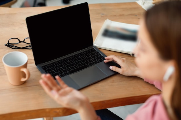 Schoolgirl using laptop with blank screen gesturing while talking at webcam sitting at desk at home having video call