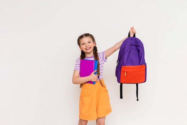 A schoolgirl with books in her hands lifts up a school backpack on a white isolated background Preparation for the academic year