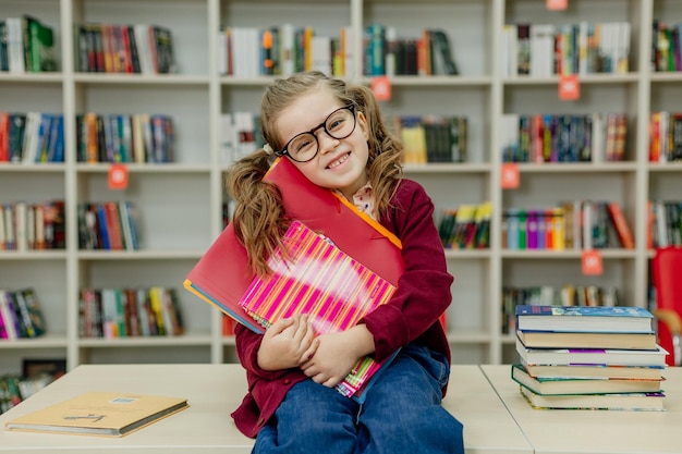 Schoolgirl with glasses is sitting at a desk with a huge stack of books textbooks in the library