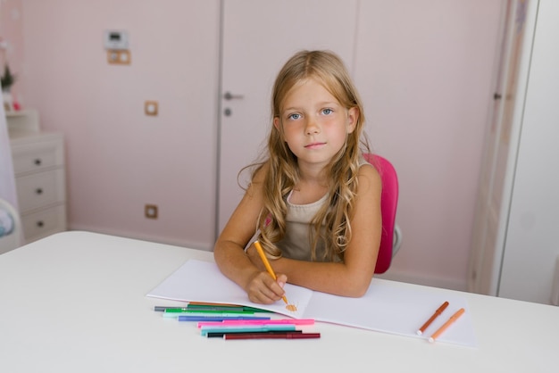 Schoolgirl with a pensively draws in a notebook with her right hand or does homework and looks at the camera