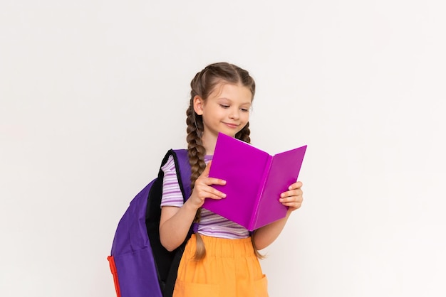 A schoolgirl with a satchel behind her reads a book on a white isolated background Summer vacation classes