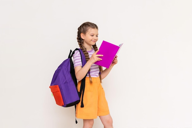 A schoolgirl with a satchel behind her reads a book on a white isolated background Summer vacation classes