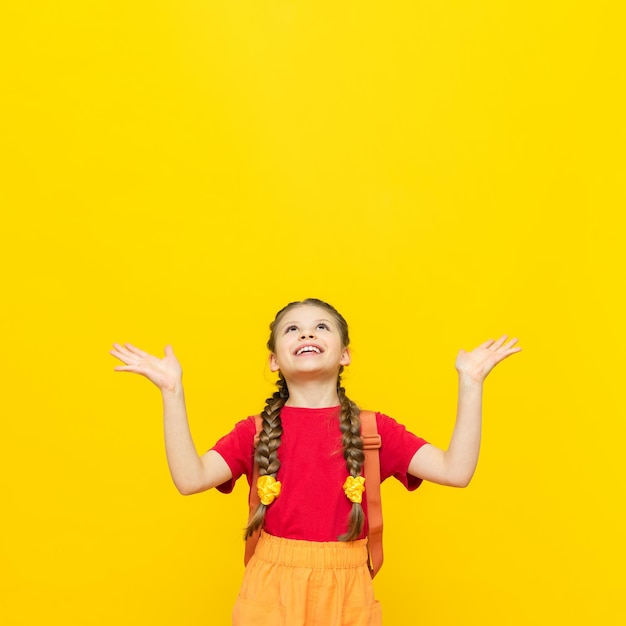 A schoolgirl with a satchel looks at your advertisement for educational courses for schoolchildren A little girl is preparing for exams on a yellow isolated background Copy space