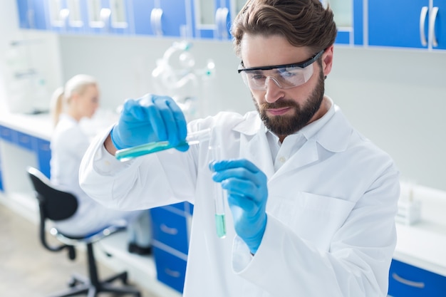 Scientific experiment. Serious nice professional chemist wearing protective glasses and pouring liquid from one test tube to another while doing scientific experiment