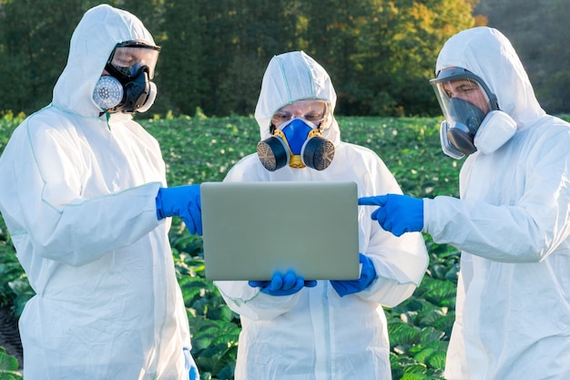 Scientist and Agronomists wearing a white protective equipment, chemical mask and glasses use Laptop on farm field.