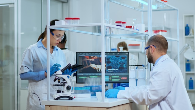 Scientist nurse taking notes on tablet in modern equipped laboratory. Multiethnic team examining vaccine evolution using high tech for research in treatment development against covid19 virus