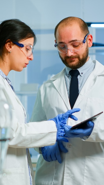 Scientist team meeting and discusssing treatment results standing in research laboratory pointing on tablet. Stuff examining vaccine development against covid19 virus using high tech for researching.