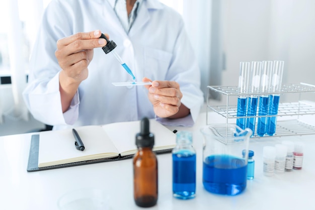 Scientist woman holding dropper to dropping solution sample on the glass plate for microscope and blue liquid in test tube while working to analyzing and developing coronavirus vaccine in laboratory