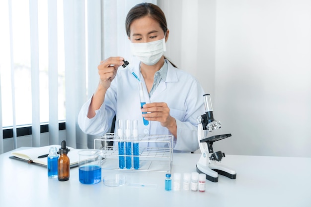 Scientist woman in protective face mask dropper to dropping blue liquid into test tube while working with microscope to analyzing and developing coronavirus vaccine in laboratory