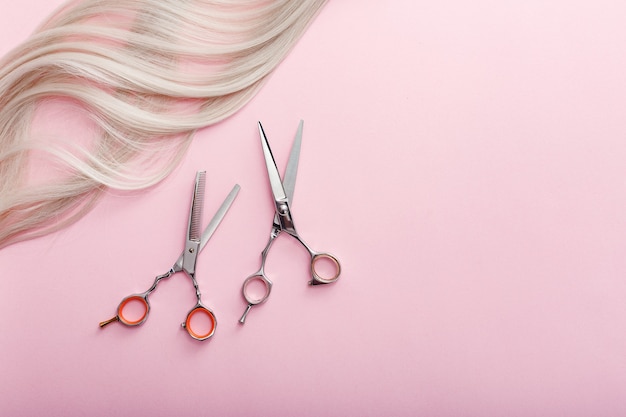 Scissors and other hairdresser's accessories and strand of blonde hair on pink background. Hairdresser service.