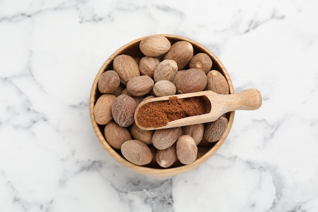 Scoop with nutmeg powder and seeds in bowl on white marble table top view