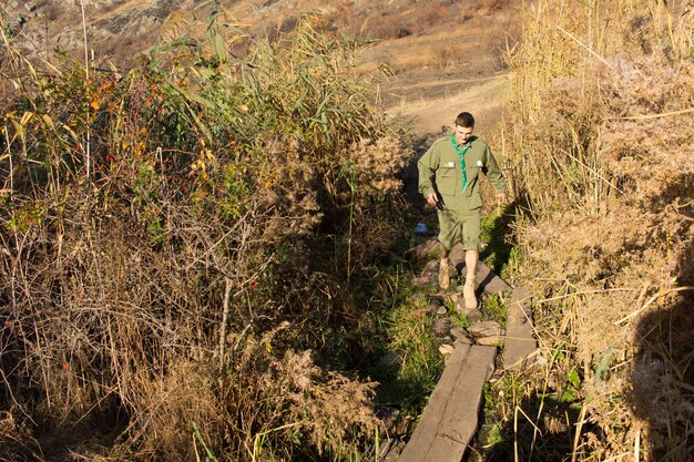 Scout crossing a narrow footbridge formed from a tree trunk over a stream or ravine as he hikes through the wilderness