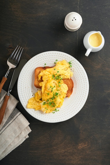 Scrambled Eggs Fluffy and Buttery scrambled eggs on bread with microgreen radish and hollandaise sauce on white plate over dark old wooden background Homemade breakfast or brunch meal Top view