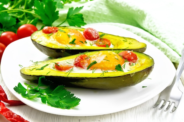 Scrambled eggs with cherry tomatoes in two halves of avocado in a plate, green towel and fork on the background of white wooden board