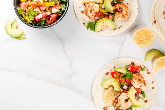 Seafood. Mexican food. Tortilla tacos with traditional homemade salsa salad, parsley, fresh lemon, avocado and grilled shrimp pawns. On a white marble . Top view 