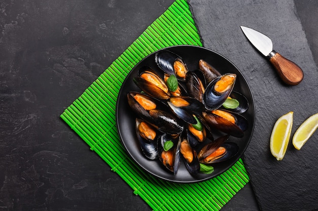 Seafood mussels, basil leaves in a black plate with lemon and knife on green bamboo mat and stone table. Top view.