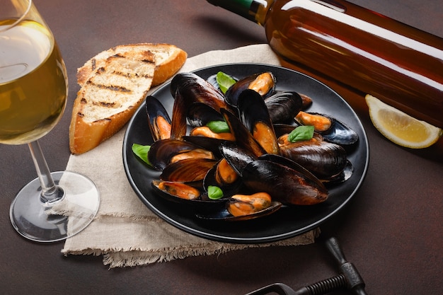 Seafood mussels and basil leaves in a black plate with wine bottle, wineglass, corkscrew, bread slices, burlap on rusty background. Top view.