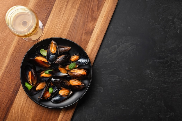 Seafood mussels and basil leaves in a black plate with wineglass, lemon, knife on wooden board and stone table