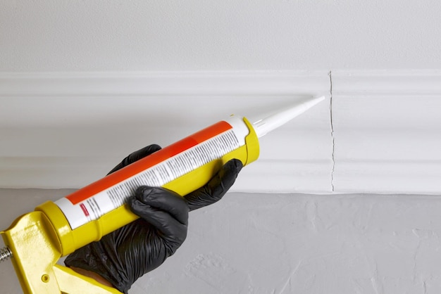 Sealing gaps in the joints of ceiling plinths Hands in black gloves hold a gun with acrylic sealant