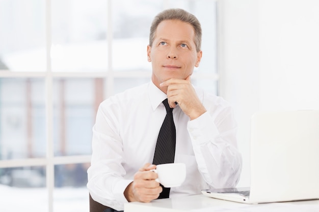 In search of the new ideas. Thoughtful mature man in formalwear sitting at his working place and drinking coffee