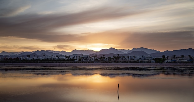 Seascape with a reflecting sky. The sun sets over the city near the mountains.