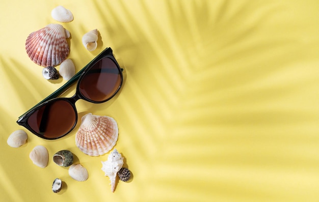 Seashells and sunglasses on a bright yellow background with palm leaf shadow The concept of summer holidays Top view and copy space