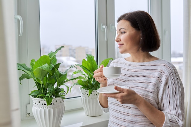 Season winter snowy day middle aged smiling woman with cup of coffee looking out the window