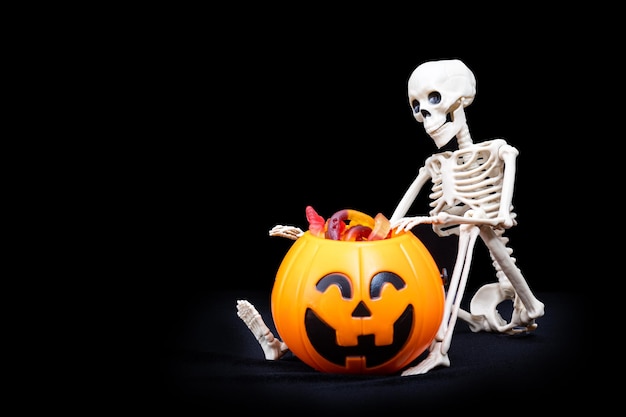 A seated skeleton pulls out of a bucket in the shape of a pumpkin candy on a black background Trick or Treat