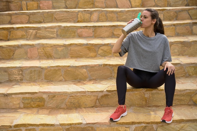 Seated sportswoman drinking water from an environmentally friendly aluminum bottle on stairs