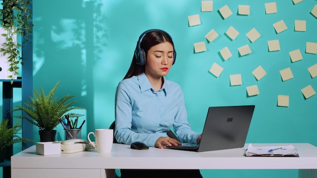 Secretary wearing headphones following directives received from manager during online videocall. Asian employee in video conference meeting with her boss in colorful office over blue studio background