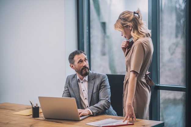 Seduce boss. Pretty blonde young woman in dress standing in front of surprised adult boss sitting at table in office