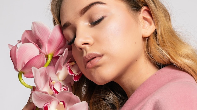 Seductive blonde woman in pink jacket posing in studio on grey background elegant model in pastel casual spring outfit Beautiful girl with healthy skin and branch orchid 169 panoramic format