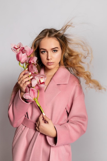 Seductive blonde woman in pink jacket posing in studio on grey background Fashion portrait of elegant model in pastel casual spring outfit Beautiful girl with healthy skin and branch orchid