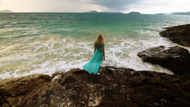 Seductive longhaired woman walks on rock of sea reef stone stormy cloudy ocean Woman in blue swimsuit dress tunic Concept rest in sea tropical resort coastline traveling tourism summer holidays