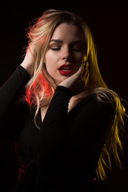 Seductive young blonde model with bright makeup and long wavy hair posing over a black background