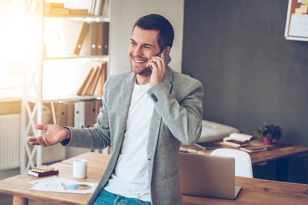 See you in my office! Handsome young man talking on mobile phone with smile while leaning to the table at his office