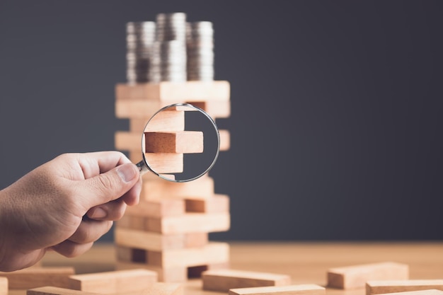 Seeking about risks in business or financial Idea to find risky in business Businessman using magnifying glass to find risk point on tower wooden block game on wooden desk in office