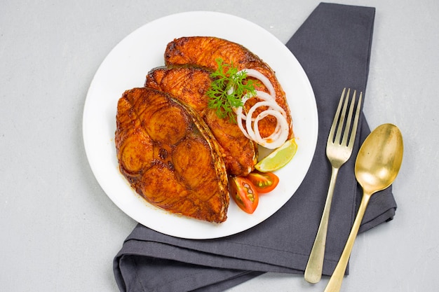 Seer fish fry arranged beautifully and garnished with onion lemon and tomato slices on white ceramic plate placed on dark grey colour napkins with grey textured background