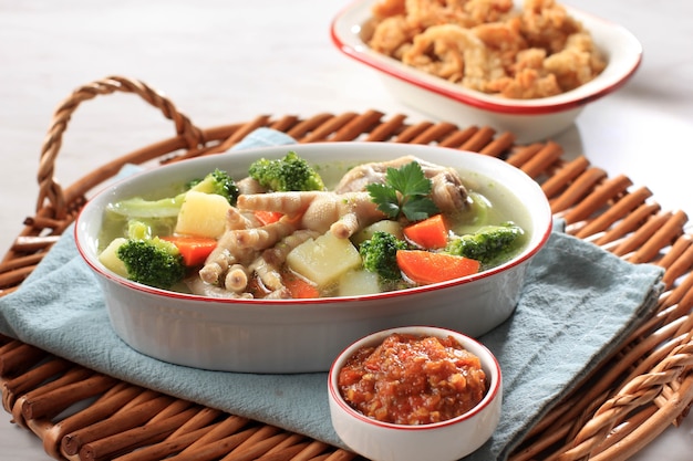 Photo selected focus chicken feet (ceker) on vegetable clear  soup with potato, broccoli, and carrots. served on wooden table in oval bowl with sambal. and oyster mushroom copy space for text