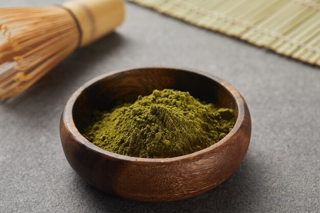 Selective focus of bamboo whisk and green matcha powder in wooden bowl on grey table with copy space