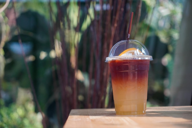 Selective focus cold Iced black coffee mixed with yuzu juice and orange slices in plastic glass on wooden table in nature background two tones layer fresh summer drink in the morning