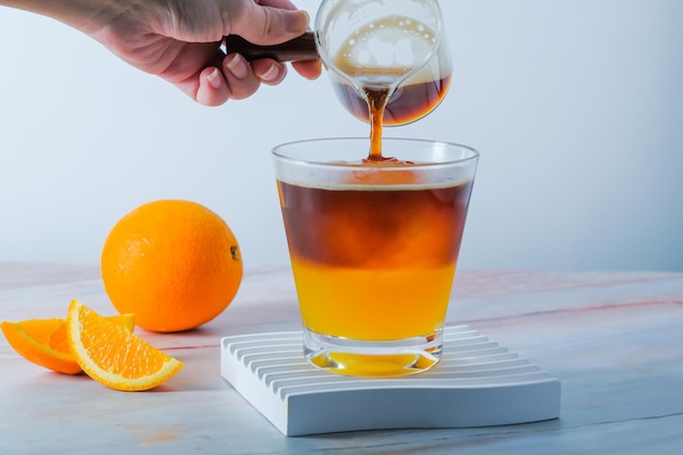 Selective focus cold Iced glass of espresso with orange juice and fresh sliced orange on wooden plate in white background Summer Cocktail Cold brew coffee or black tea Pouring cold drink