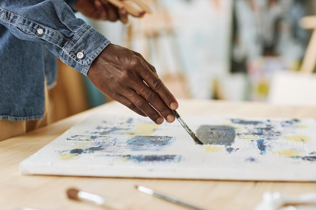 Selective focus on hand of young black man painting on white canvas with acrylic paints while standi