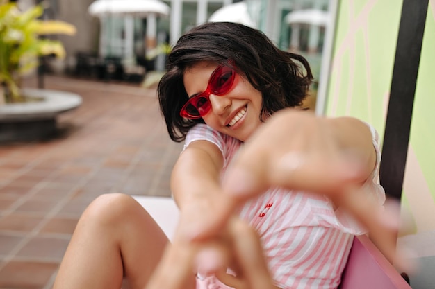 Selective focus of laughing brunette lady with outstretched hands Outdoor shot of blithesome girl in pink sunglasses on city background