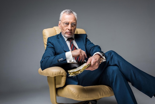 Selective focus of mature businessman sitting in armchair and holding dollar banknotes on grey