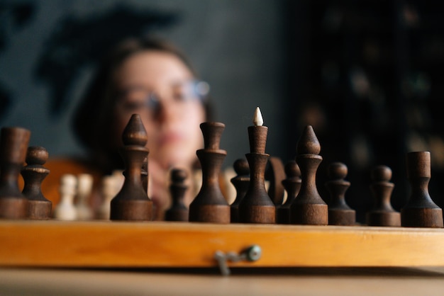 Selective focus of pensive young woman wearing elegant eyeglasses thinking about chess move while sitting on wooden floor in dark room