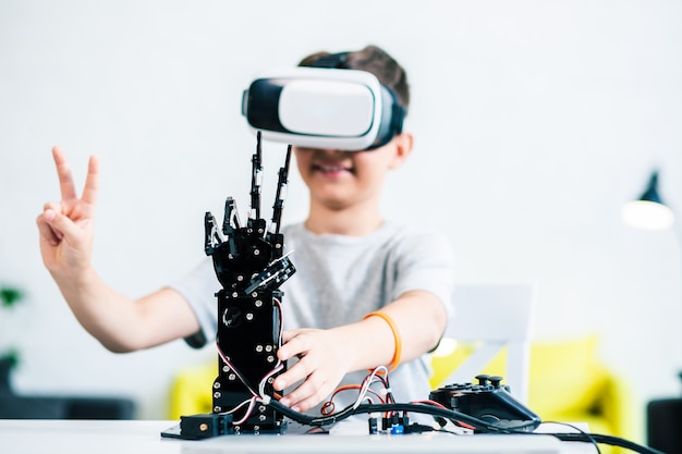 Selective focus of a robotic hand standing on the table while a positive ingenious boy experimenting with it